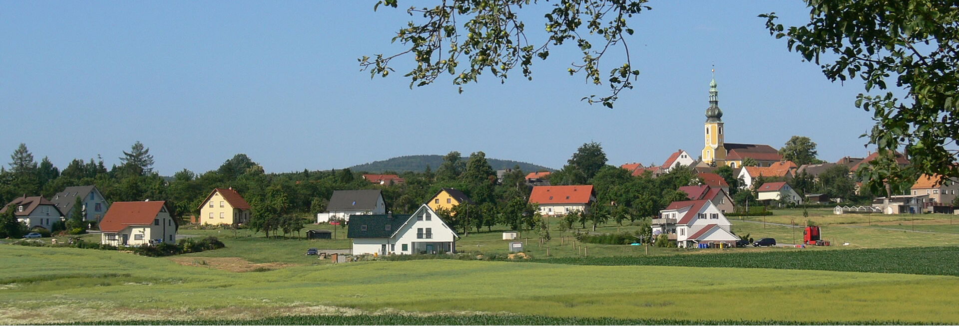 Weitblick auf die Gemeinde Hochkirch mit Feldern, Häusern und der markanten gelben Kirche unter blauem Himmel.
