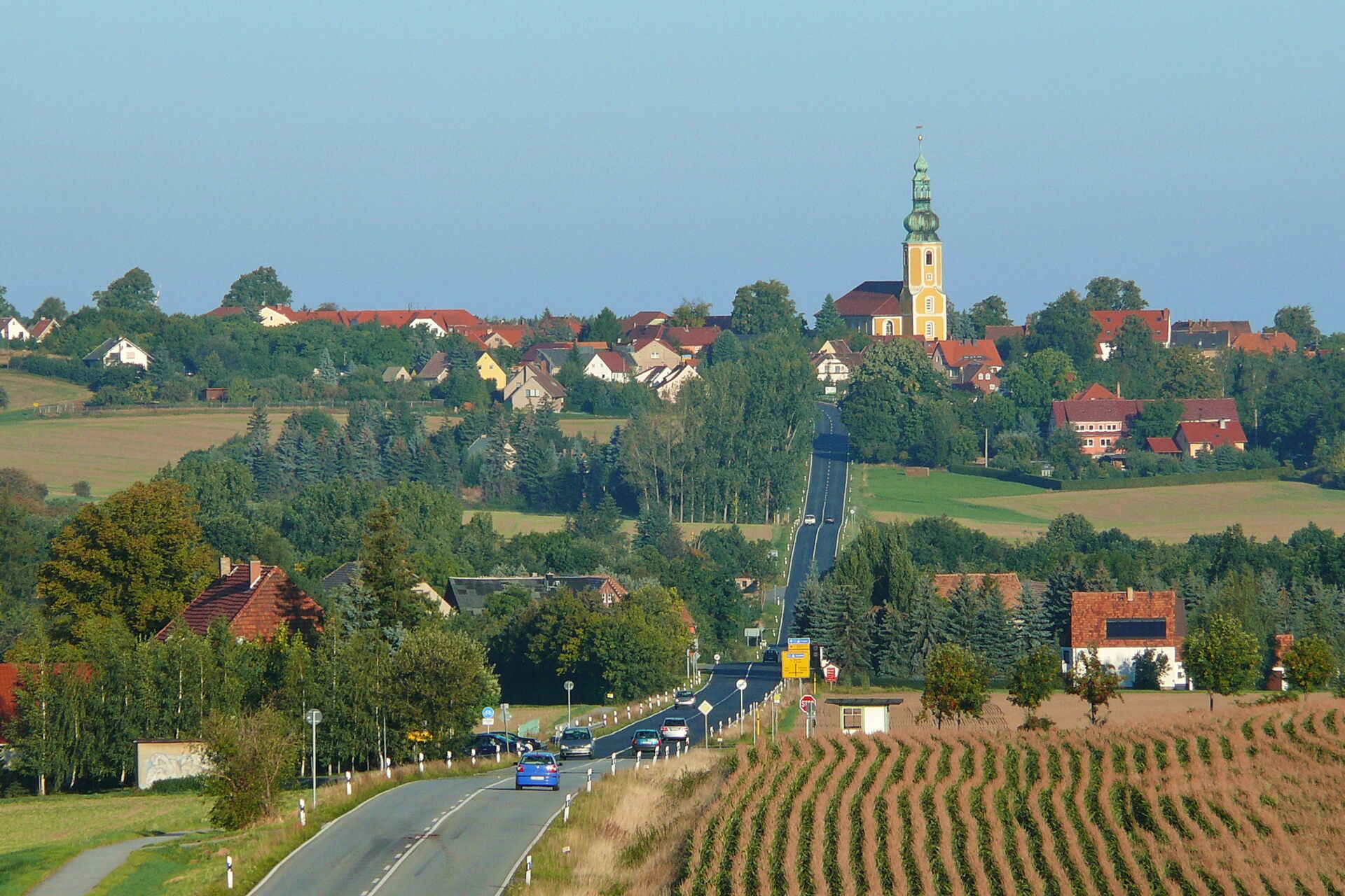 Weitblick auf die Gemeinde Hochkirch mit einer Straße, Feldern und der markanten gelben Kirche auf dem Hügel.