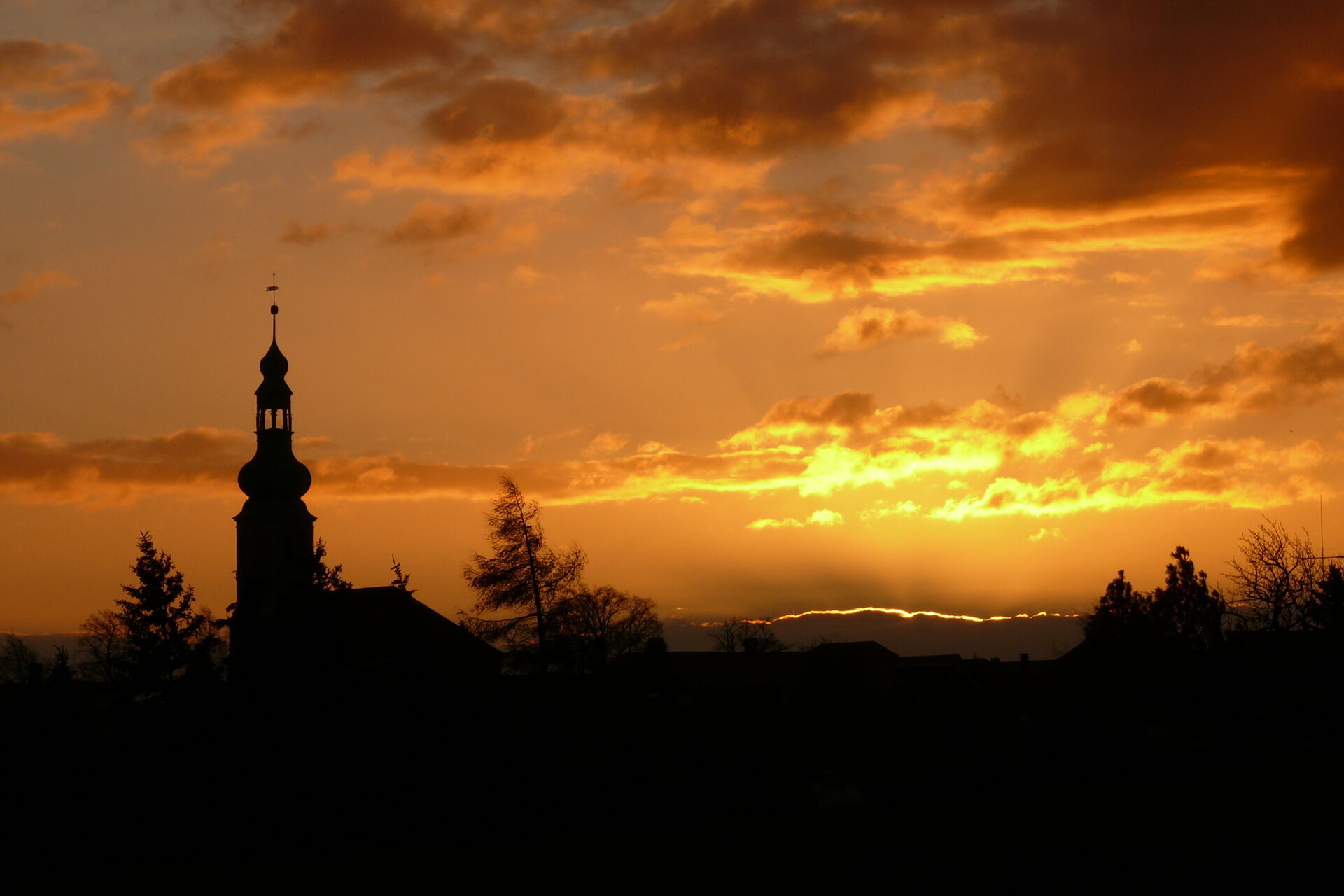 Silhouette der Kirche Hochkirch und Bäume vor einem dramatischen Sonnenuntergang mit orangefarbenem Himmel.
