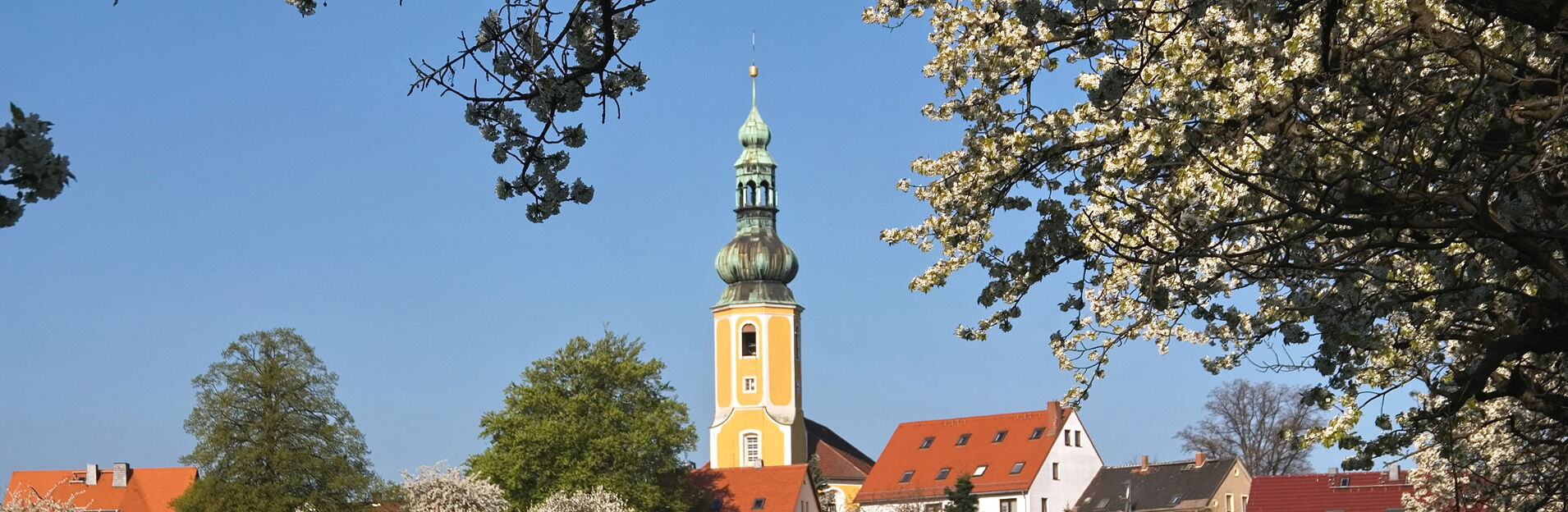 Malerische Frühlingsansicht von Hochkirch mit blühenden Bäumen, grünen Feldern und der gelben Dorfkirche unter blauem Himmel.