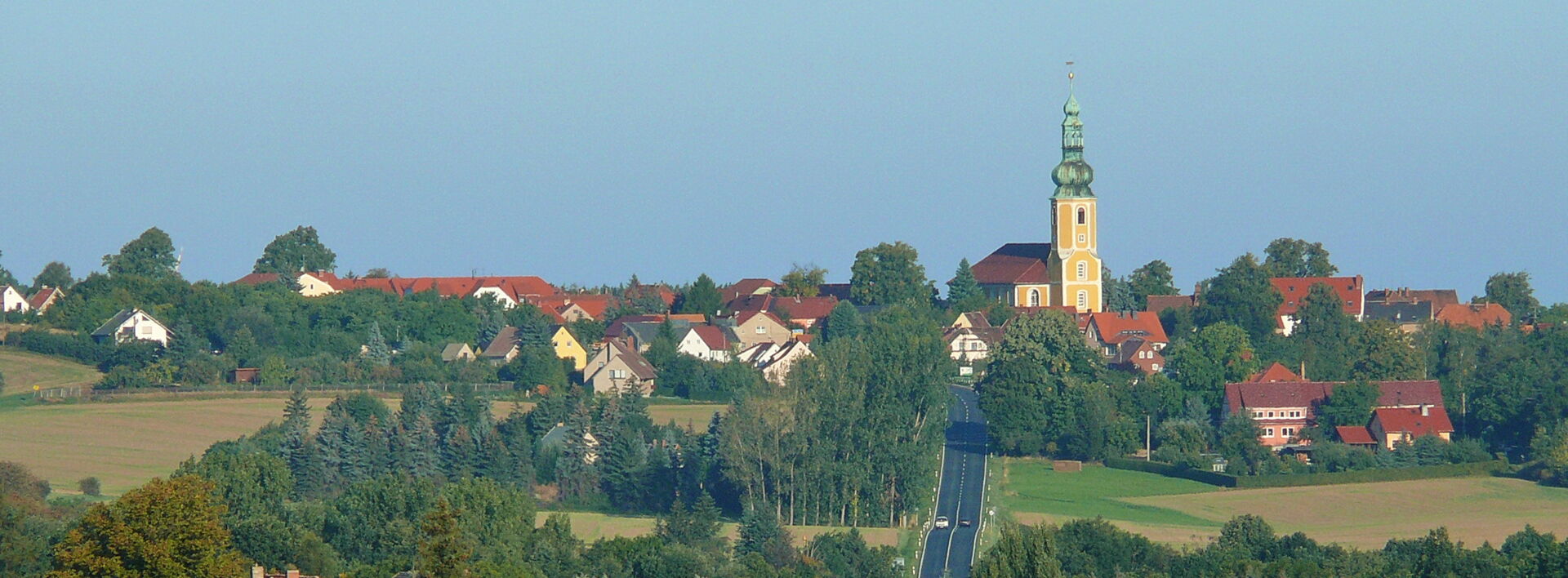 Weitblick auf die Gemeinde Hochkirch mit einer Straße, Feldern und der markanten gelben Kirche auf dem Hügel.